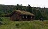 Liten trehytte med torvtak på en grønn eng i landskap med ås i bakgrunnen.