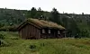 Liten trehytte med torvtak på en grønn eng i landskap med ås i bakgrunnen.