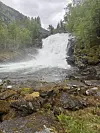 En kraftig foss faller ned i et skogkledd fjellområde med stein i forgrunnen.