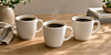 Tre hvite kaffekopper står på et lyst trebord med bøker og grønne planter i bakgrunnen.