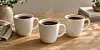 Tre hvite kaffekopper står på et lyst trebord med bøker og grønne planter i bakgrunnen.