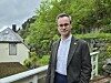 En mann i jakke står ved et rekkverk foran et hus og grønn skogkledd bakgrunn.