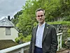 En mann i jakke står ved et rekkverk foran et hus og grønn skogkledd bakgrunn.
