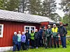Flere personer står på gress foran en rød hytte med hvitt tak og skog bak.