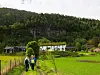 Flere personer går på en sti forbi jorder og hus med en skogkledd ås bak.