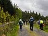 Folk med ryggsekker går på en grusvei mellom skog og gjerde under gråvær.