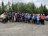 En gruppe mennesker står samlet på en grusplass foran en tett skog, flere holder turstaver.