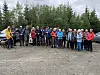 En gruppe mennesker står samlet på en grusplass foran en tett skog, flere holder turstaver.