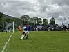 Fotballspillere i kamp på en gressbane foran mål og tribuner under overskyet vær.