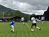 Fotballspillere spiller på en gressbane foran fjell og tribune.