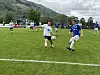 Fotballspillere kjemper om ballen på en grønn bane med fjell i bakgrunnen.