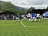 Fotballspillere på en gressbane med tribune og skog i bakgrunnen.