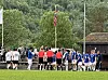 To fotballag står samlet på en gressbane foran skog og en liten bygning.