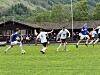 Fotballspillere i kamp på en gressbane med tilskuere og fjellskog i bakgrunnen.