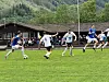 Fotballspillere i kamp på en gressbane med tilskuere og fjellskog i bakgrunnen.
