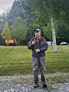Mann med fiskestang står på et gressområde med trær og fjell i bakgrunnen.