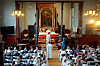 En fullsatt kirke med mennesker sittende i benker mens presten ber for konfirmantene, en og en.