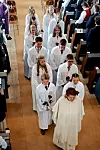 En prest og flere konfirmanter i hvite kapper går i prosesjon i en kirke med publikum langs midtgangen.