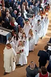En prest og flere konfirmanter i hvite kapper går i prosesjon i en kirke med publikum langs midtgangen.