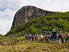 Mange turgåere står samlet på en eng foran en bratt fjellvegg og grønn skog.
