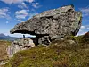 Stor steinformasjon med overheng i fjellandskap og en liten person ved foten.