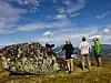 Tre personer står på en steinete fjellknatt og ser utover et vidstrakt landskap under blå himmel.