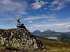 Person sitter på en fjellknaus og ser utover fjell, innsjøer og et åpent landskap under blå himmel.