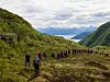 Mange turgåere på rekke på en sti i et grønt fjellandskap med fjord og fjell i bakgrunnen.