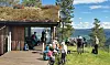 Folk står og sitter ved en fjellhytte med utsikt over innsjø og skogkledd natur.