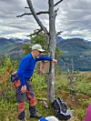 En person i blå jakke står ved et tre på en fjelltopp med utsikt over landskapet.
