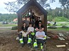 En gruppe personer sitter og står foran en liten trehytte i et grønt parkområde.