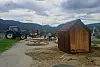 Landlig område med små trebygninger, traktor og steinbenker foran fjell og overskyet himmel.