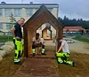 Tre arbeidere i fluorescerende arbeidsbukser ved en liten trekonstruksjon utendørs.