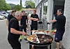 Menn står rundt en stor grill ved en bygning mens mat steker og et bord med utstyr står ved siden av.