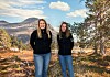To kvinner står foran et stort bilde av fjell, vann og høstfargede trær.