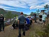 Personer står og sitter ved et utsiktspunkt med fjellandskap, vann og skyet himmel.