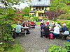 Flere personer sitter ved picnicbord i en frodig hage foran et gult hus.