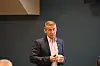 Mann i dressjakke holder en kopp foran en mørk vegg i et konferanserom.