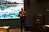 En foredragsholder står foran en presentasjon med bilde av is og fjell i en konferansesal.