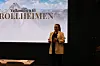 En dame står på en scene foran en skjerm med teksten «Velkommen til Trollheimen».
