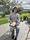 Jente sitter på en stol på en treterrasse og karder ull, med grønt landskap og fjord bak.
