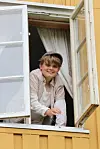 Ung person ser ut og smiler fra et åpent vindu på en gul bygning med hvite vindusrammer.