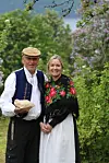 To personer står sammen i en grønn hage, iført tradisjonelle klær med fargerike broderier.