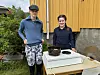 To personer står ved en gryte på en liten utendørs komfyr foran en gul vegg.