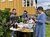 Tre personer står bak to bord med mat og drikke foran en gul bygning i en hage. En dame står på andre siden av et av bordene og ser på en flaske.