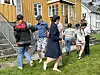 Flere personer danser på gress ved siden av en gul bolig med trapper og hvite vinduer.