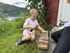 Et gutt sitter på gresset og hammer på en spiker som står i en treklosse(r) ved en rød bygning og en større gutt.
