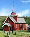 Rød trekirke med høy spire, hvite detaljer og grønt gress foran skog og blå himmel.