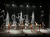 Dansere opptrer på en opplyst teaterscene med svarte gardiner og abstrakte hvite lysmønstre i bakgrunnen.