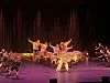 Et danseensemble i fargerike kostymer opptrer på en scenen med mørk bakgrunn og scenelys.